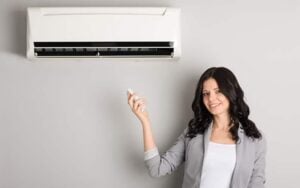 Woman in business casual attire holding remote control while standing beneath a wall-mounted ductless mini split air conditioning unit.